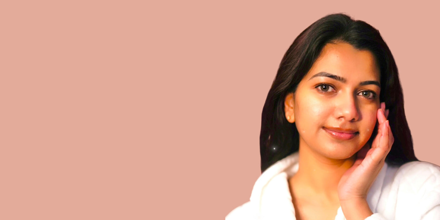 A young Indian woman with radiant, glowing skin gently touching her face, wearing a white robe against a soft beige background, representing natural beauty and skincare.